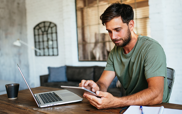 man sitting at desk looking at notepad with laptop on desk